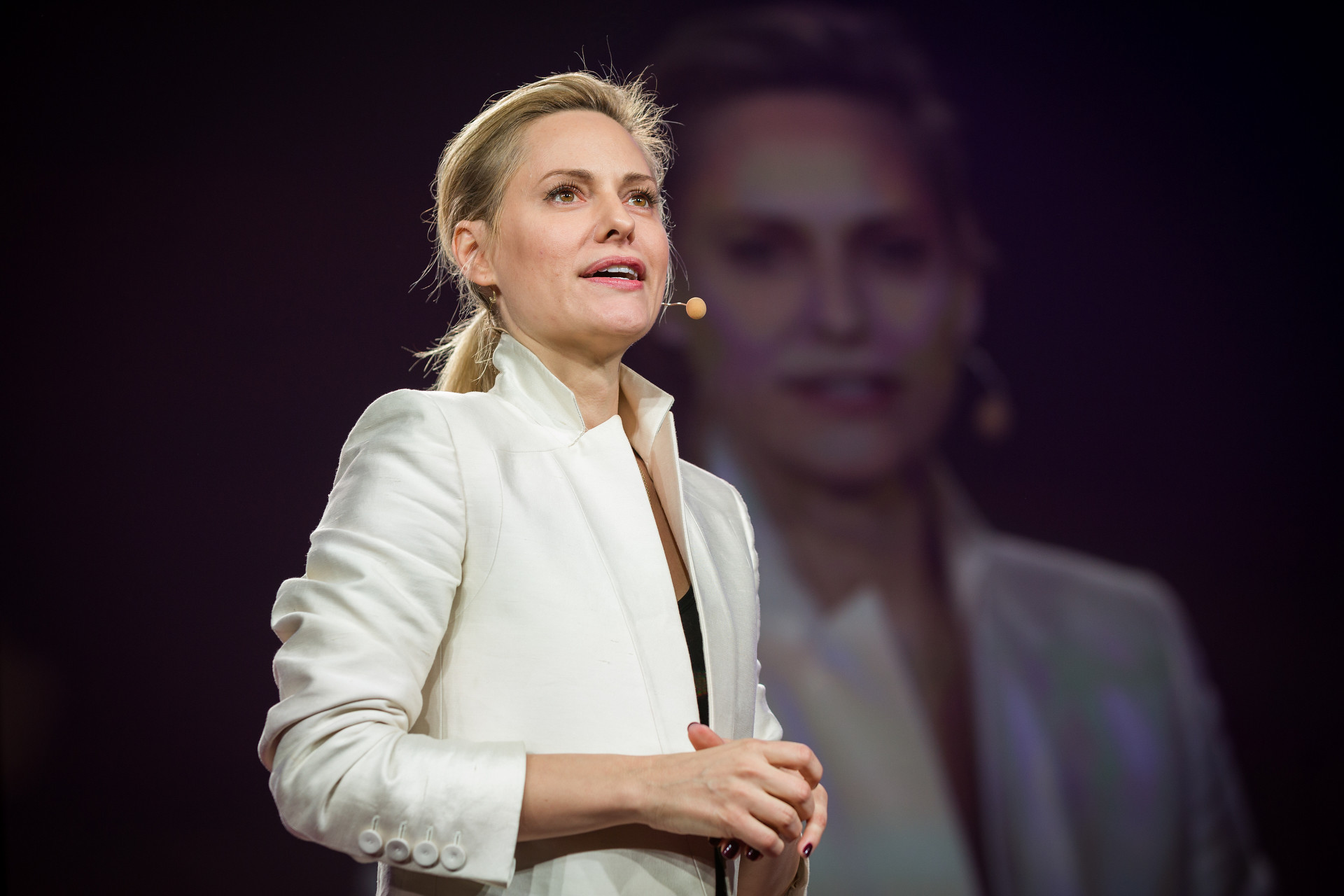 "TED2014_RL_2R9B9133_1920" by TED Conference is licensed under CC BY-NC 2.0. Image shows Aimee Mullins wearing a white jacket with a a white microphone headset on, standing in front of a photo or video or herself on a large screen