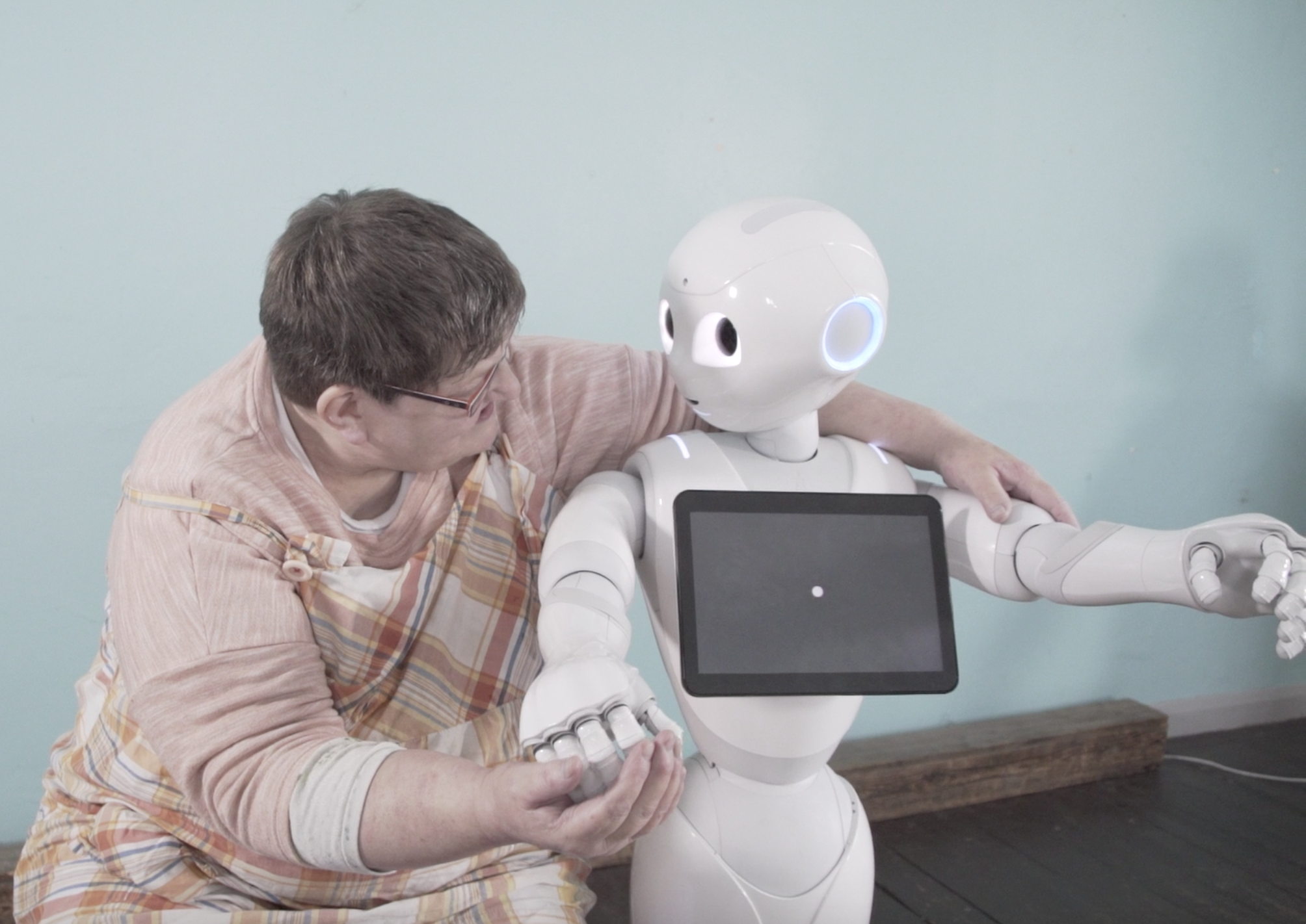 a woman puts her arms around a white robot figure in the pose of a hug. The robot has its head turned to look at her face. The robot has a tablet screen across its chest