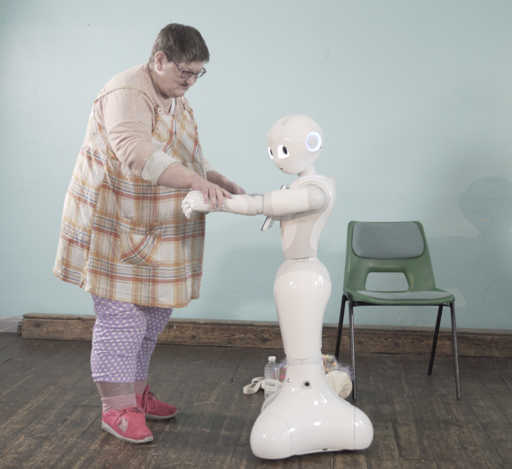 a woman standing in a room holds both hands of a white robot as if they are about to dance, facing one another