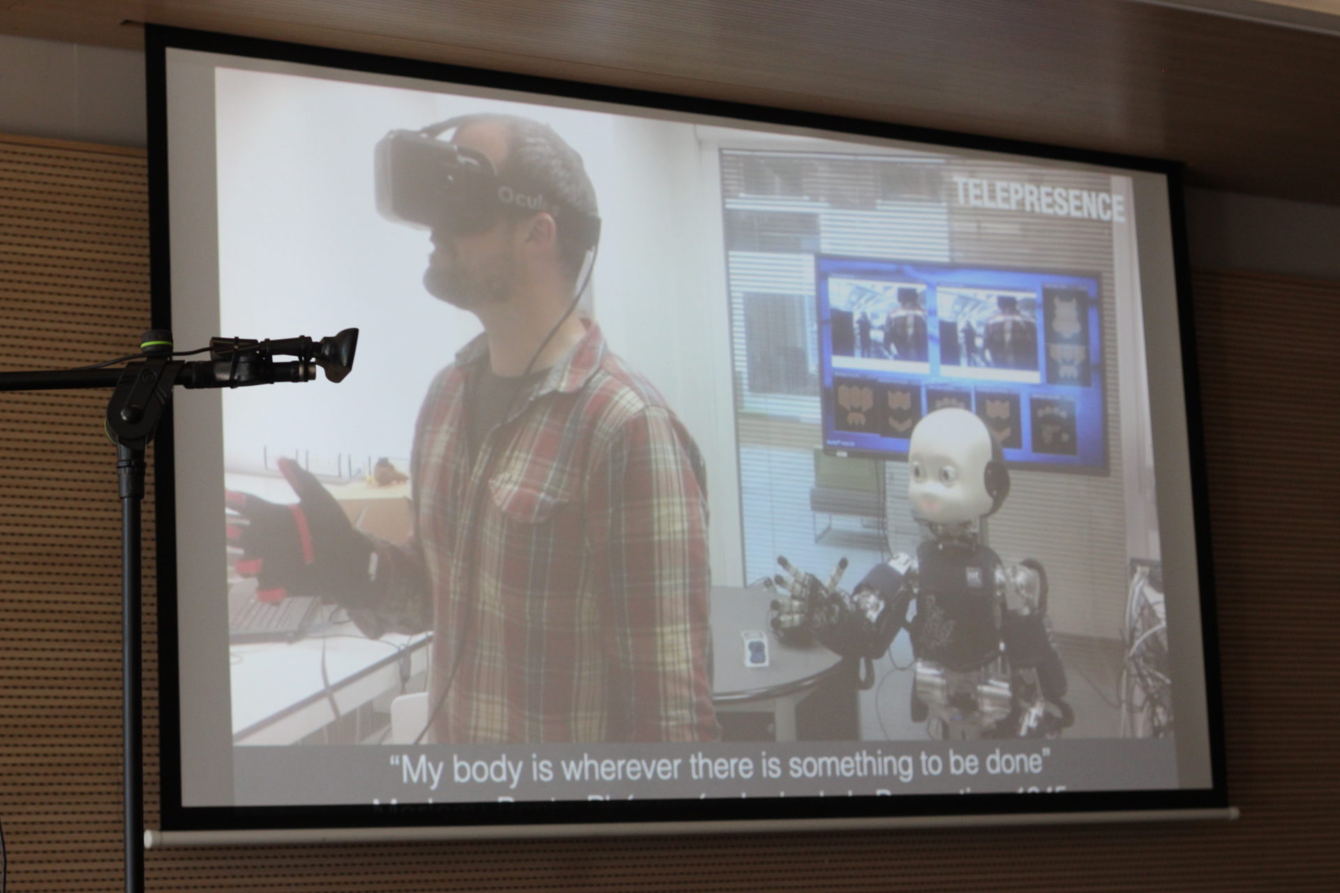 A PowerPoint slide on a big screen of a man using a VR headset to teleoperate a small, humanoid robot.