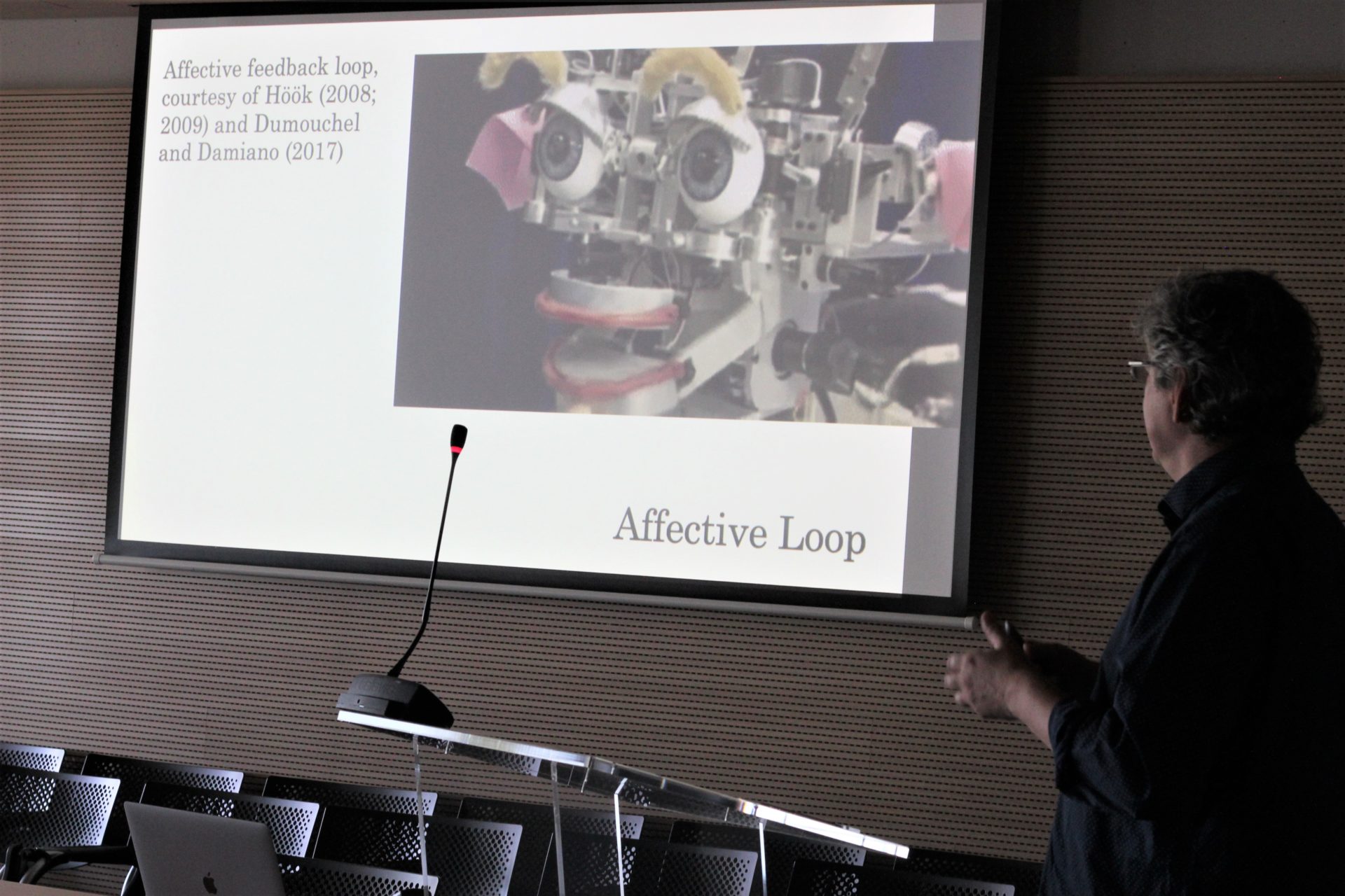Mark Paterson stands to the side of a PowerPoint slide on a big screen showing affective feedback loops, featuring a humanoid robot’s face stripped of its “skin”.