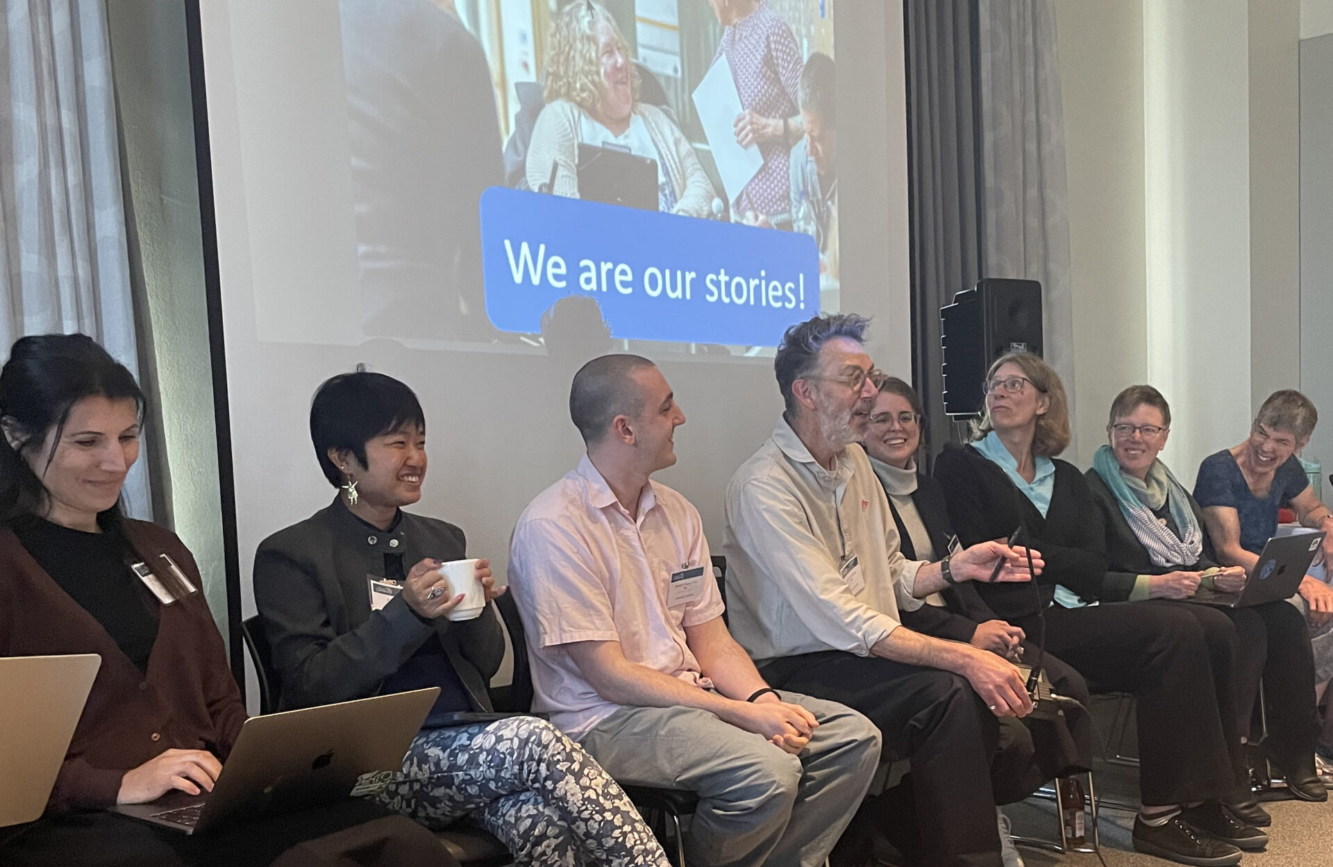Eight people – a mix of female and male; disabled and non-disabled; of younger and older working ages – are seated in a row, enjoying a convivial discussion. A screen above shows an image of disabled and non-disabled people in conversation, with a large title 'We are our stories!'