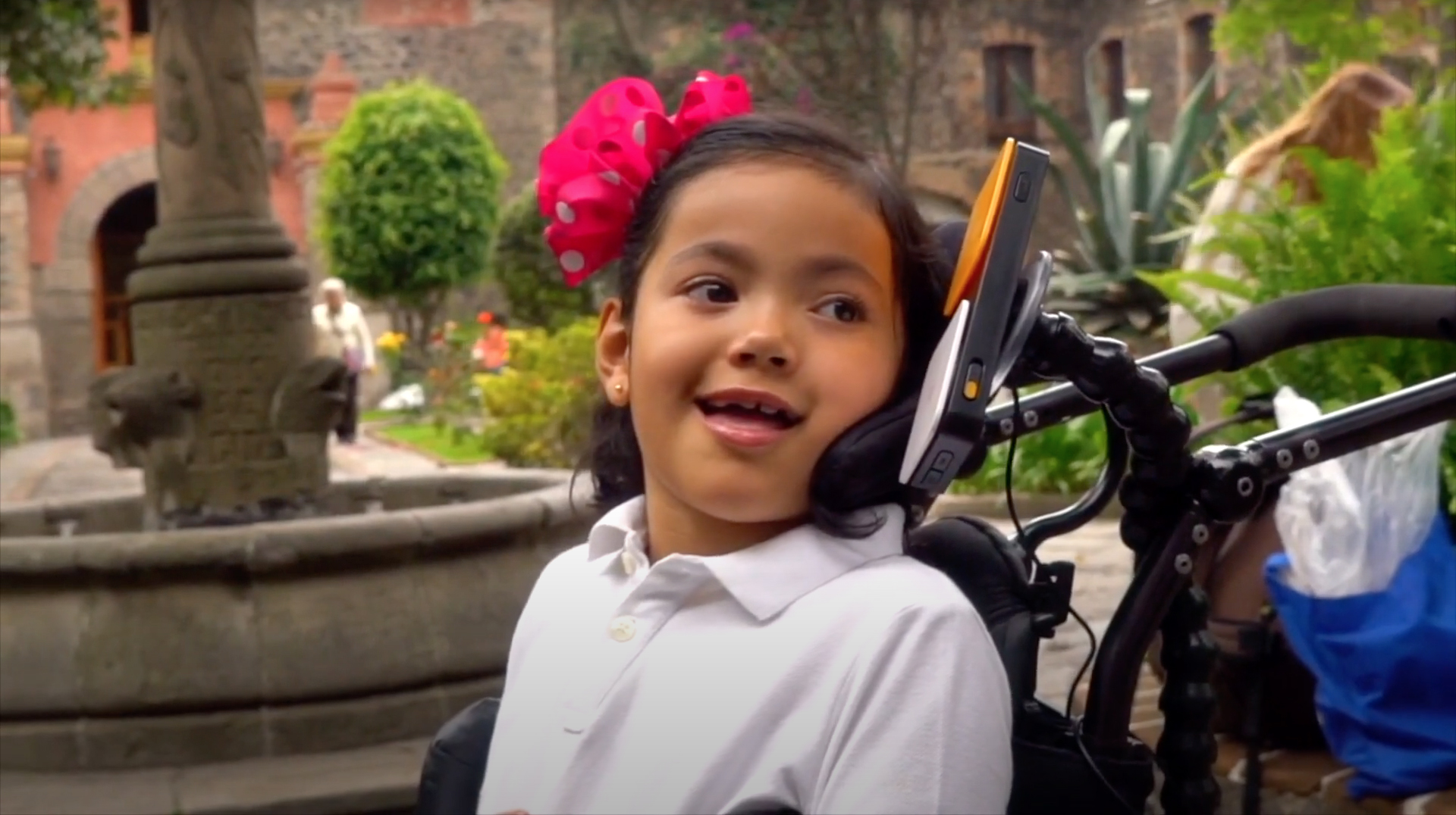 A young girl is smiling at something or someone to the right of the camera. Her hair is tied with a magenta ribbon with white polka dots, set off by the lush greens of the tropical plants in a courtyard garden. She has two large switches, one yellow, one white, on the headrest of her wheelchair. Other objects of daily life include some polythene bags on a low wall behind her.