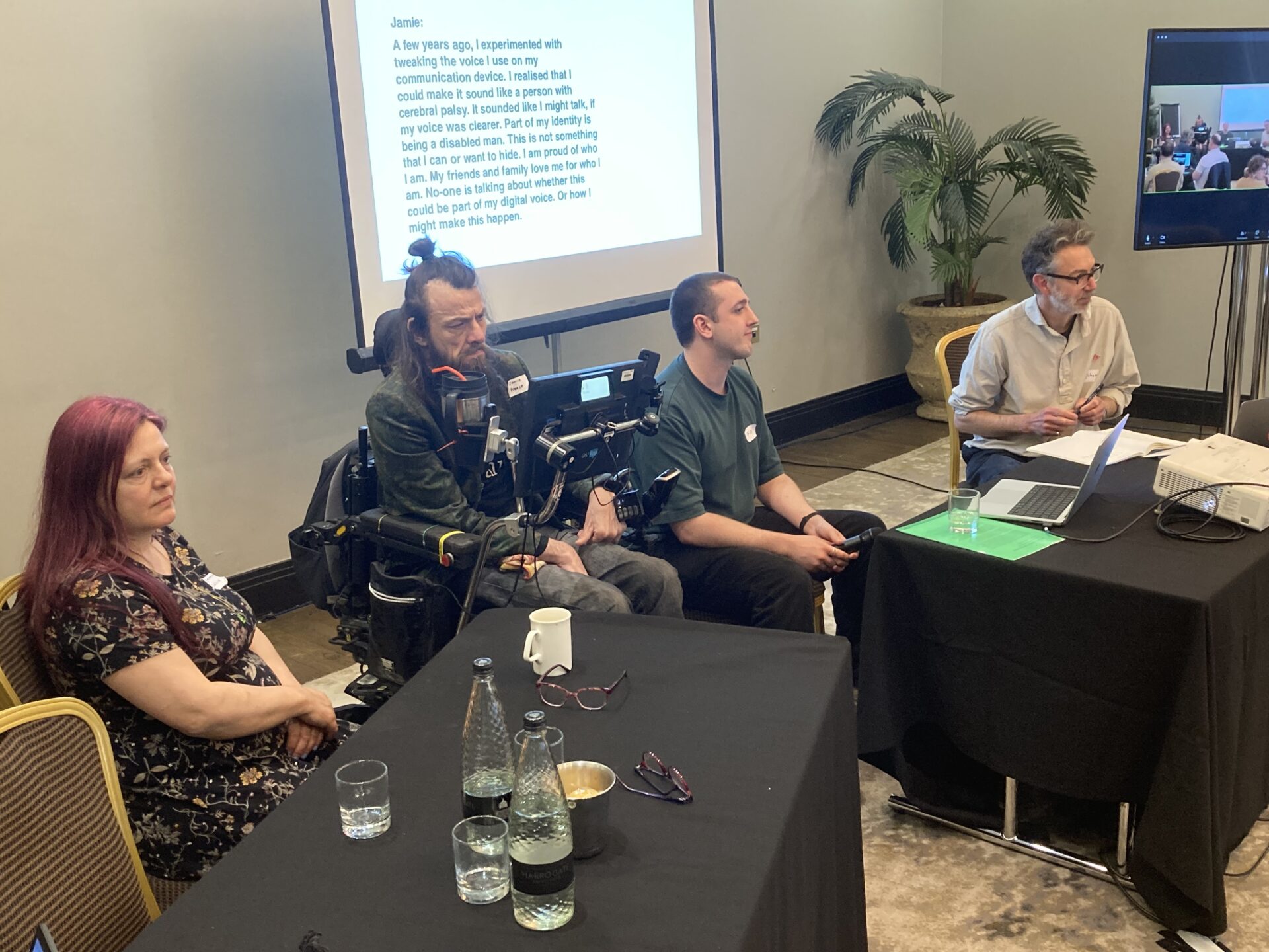 Four people – one in a wheelchair – sitting behind two desks and speaking to a conference audience with a screen behind them displaying a PowerPoint slide.