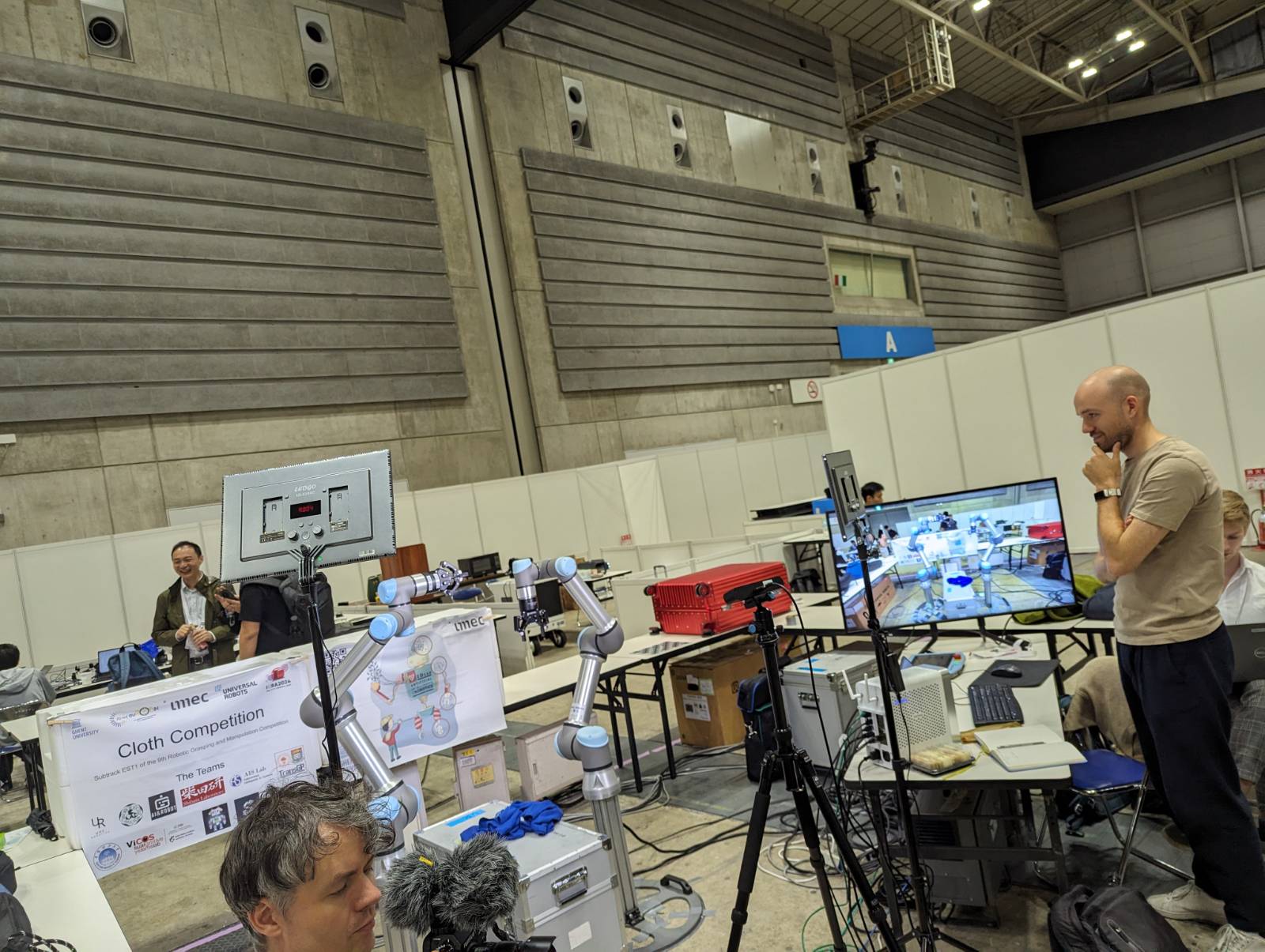 Robotic arms programmed by Shibata Lab students take part in a clothes folding competition at the International Conference on Robotics and Automation (ICRA). Held inside a large exhibition hall, there are a number of tables, an LCD screen, crates and boxes, a mass of electrical wiring, computers and 6 people can be glimpsed behind various pieces of equipment