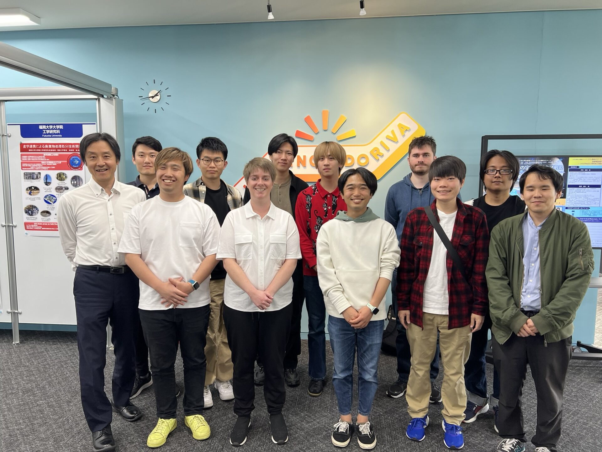 A group photo of people smiling to the camera. Christina Stimson with Shibata-sensei and his 10 students, standing in front of a blue wall. To the rear can be seen some academic posters.